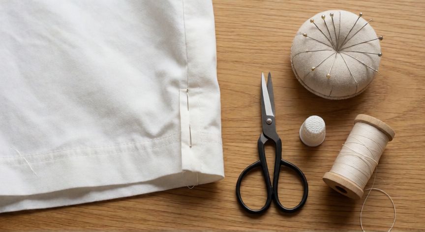 como coser invisible a mano, Mesa de trabajo de costura con dobladillo sujeto con alfileres, tijeras, dedal e hilo sobre superficie de madera