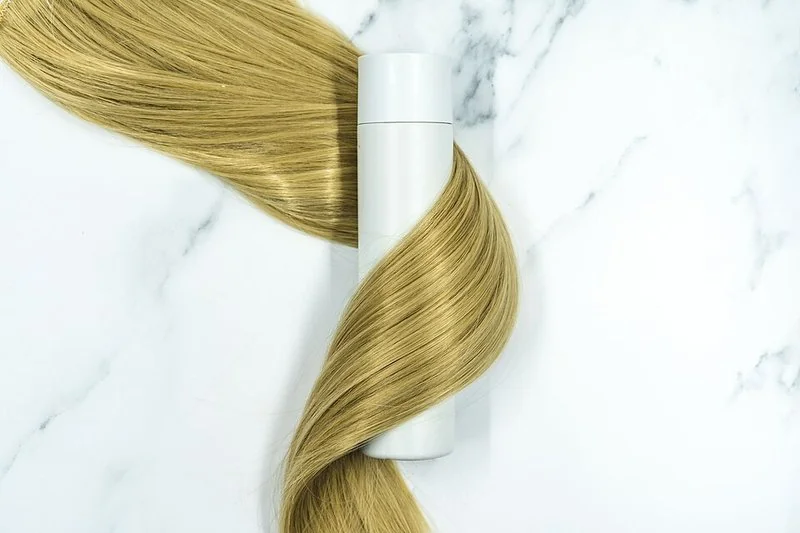 mejor producto para el pelo, Mechón de cabello rubio largo y brillante enrollado en un envase blanco de producto capilar
