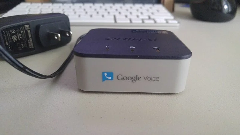 Dispositivo OBi con el logotipo de Google Voice sobre un escritorio junto a teclado y cargador