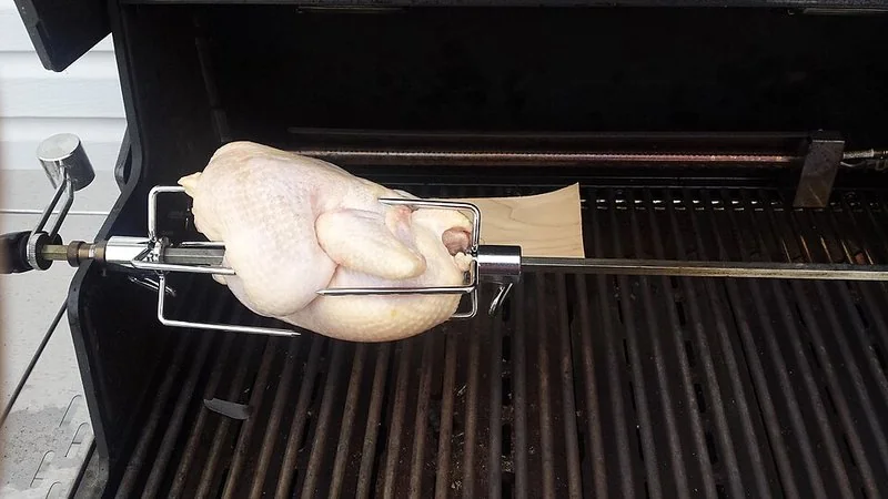Pollo entero ensartado en un espetón giratorio sobre la parrilla, listo para asarse dando vueltas