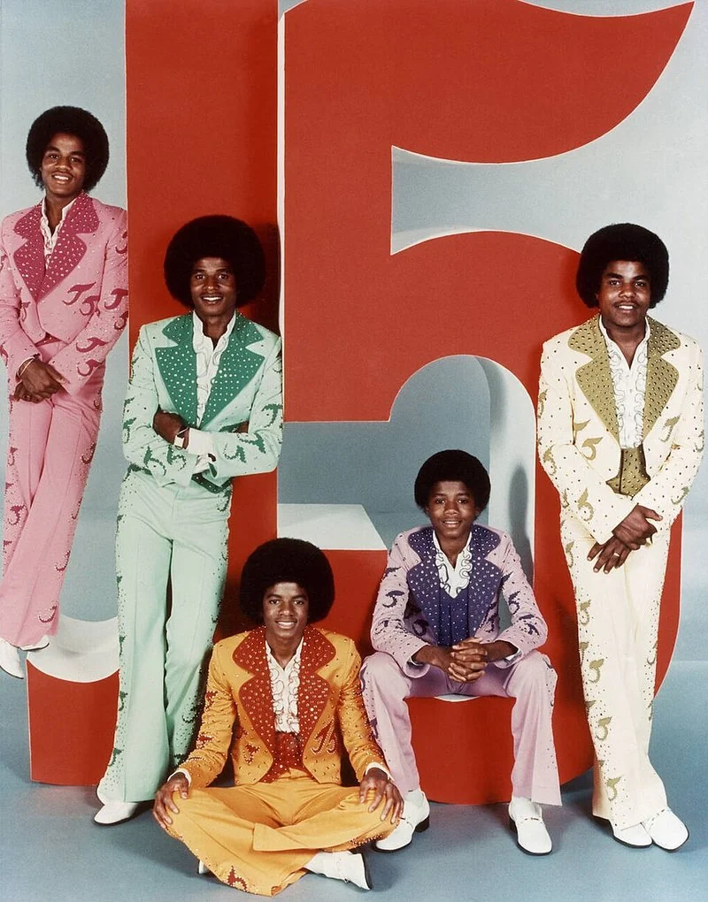 que hicieron los hermanos jackson, Retrato promocional en color de The Jackson 5 con trajes de colores y melenas afro delante del número 5 rojo