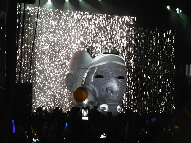 Avicii actuando en directo en el festival Lollapalooza de 2012, con una enorme máscara luminosa sobre el escenario y cascadas de luces sobre el público