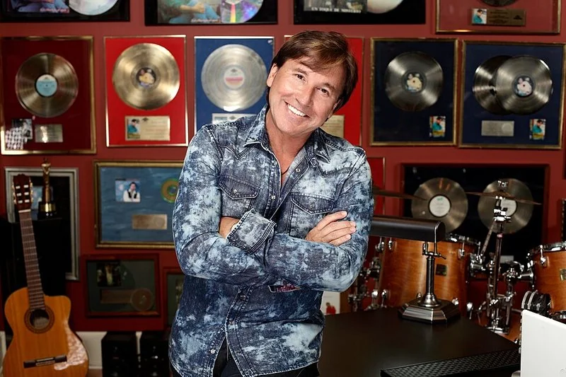 Ricardo Montaner sonriente, de brazos cruzados, en un estudio rodeado de discos de oro y platino