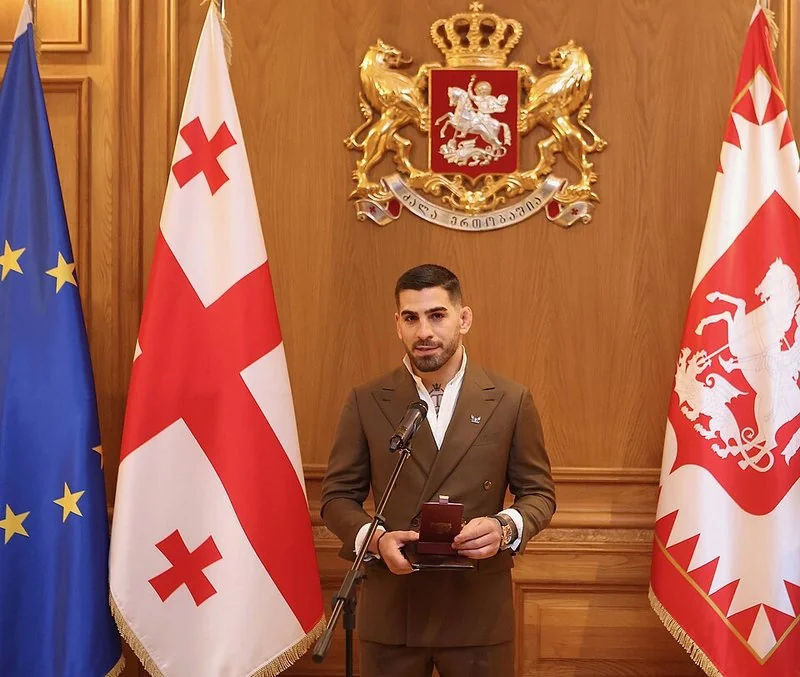 Ilia Topuria, de traje marrón, recibe una condecoración oficial ante el escudo y las banderas de Georgia
