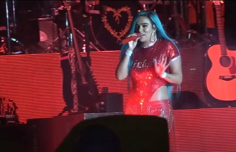 Karol G actuando en el escenario con el Bichota Tour, vestido rojo y micrófono en mano