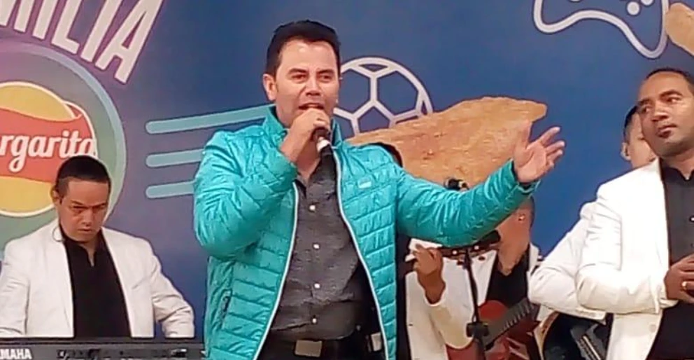 Jhonny Rivera cantando en concierto con su banda al fondo, vestido con una chaqueta acolchada turquesa y sujetando el micrófono (fichero File:JhonnyRivera.jpg d